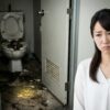 汚れていて入れないトイレの前に立ち尽くし、困惑の表情を浮かべる日本人女性。夢占いにおける「汚いトイレ」と「用を足せない」という葛藤、および足元にある金貨が示す金運の予兆を表現したアイキャッチ画像。